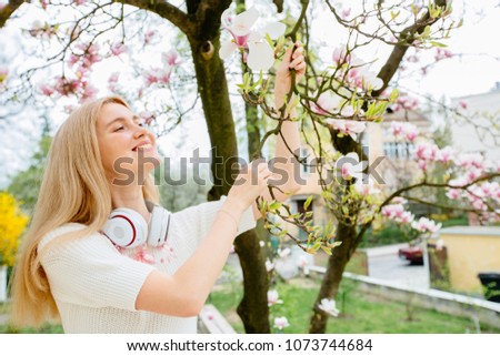 Excited teenager woman with headphones on her neck holding branch of magnolia tree and enjoying aroma of blooming garden.