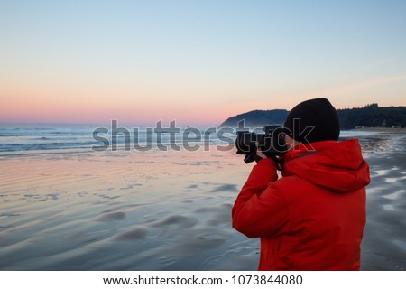Photographer with a camera is standing on the sandy beach during a vibrant and colorful winter sunrise. Taken in Canon Beach, Oregon Coast, United States of America. Royalty-Free Stock Photo #1073844080