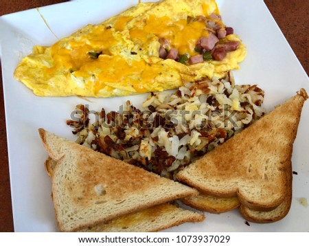 Colorful delicious photo of omelette with cheese, vegetables and ham, hash browns and white toasts. Closeup picture of homemade breakfast served on white plate on wooden table