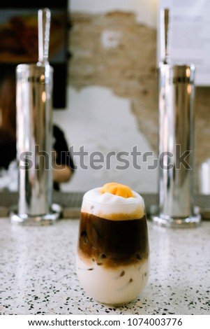 A cup of coffee topped with Thai sweet dessert and Tap in depth of field behind.