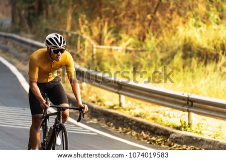 Asian man in yellow cycling jersey rinding on road bike up high on hill,  In a good weather morning, under morning sunlight. Royalty-Free Stock Photo #1074019583