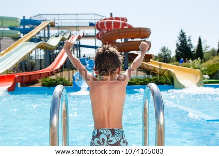 healthy happy little kid near blue swimming pool in water park with water tube slides in summer Royalty-Free Stock Photo #1074105083