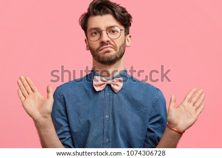 I am not guilty! Fashionable bearded guy being hesitant or shows his non participation in something, has bristle, wears stylish denim shirt, isolated over pink studio background. Body language Royalty-Free Stock Photo #1074306728