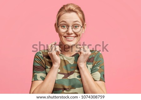 Glad young attractive cute blonde woman with blue eyes, has healthy skin, wears casual t shirt, has amazed expression, delighted to recieve positive news, isolated over pink studio background