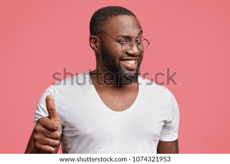 Photo of cheerful delighted dark skinned guy raises thumb, demonstrates ok sign, likes something, has shining pleasant smile, looks with cunning positive expression at camera through spectacles