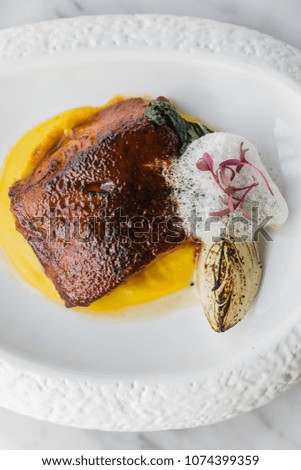 Close up of pan seared trout. served with yellow sauce, foam and roasted sliced onion on curved white plate. 