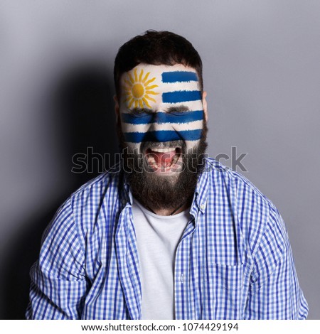 Face of young screaming man painted with flag of Uruguay. Football or soccer team fan, sport event, faceart and patriotism concept. Studio shot at gray background, copy space