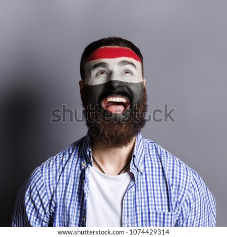 Face of young happy bearded man painted with flag of Egypt. Football or soccer team fan, sport event, faceart and patriotism concept. Studio shot at gray background, copy space