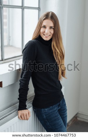 Portrait of young cheerful long-haired woman wearing black roll neck jumper standing by window