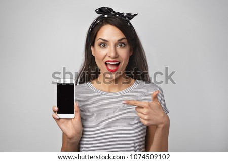 Picture of glad fascinated young woman screaming in excitement, keeping mouth wide opened, feeling happy, demonstrating her new mobile phone, pointing index finger at blank copy space screen