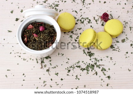 Small white porcelain pot fill of aromatic green tea with some petals and buds of pink rose and some french cakes on white wooden background. Concept for healthy food theme. Overhead view. Copy space