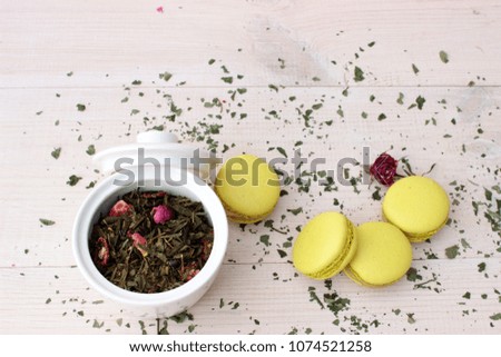 Small white porcelain pot fill of aromatic green tea with some petals and buds of pink rose and some french cakes on white wooden background. Concept for healthy food theme. Overhead view
