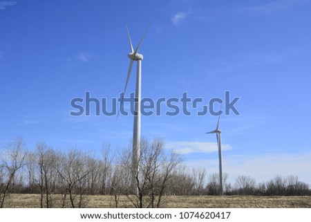 Two windmills making electricity on a beautiful blue sky spring day with some trees in the foreground and a few scattered clouds. 