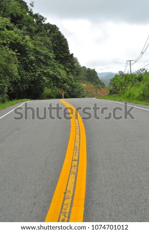 Infinity yellow lines running parallel on a local road with green trees and hills all along in Nan Province with a curved turning ahead.