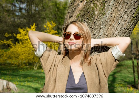 Young redhead girl in sunglasses standing near a tree in springtime season park