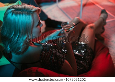 Blonde woman holding glass with cocktail while sitting in beanbag 