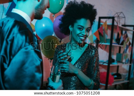 African american woman holding glass with cocktail and talking to man at party