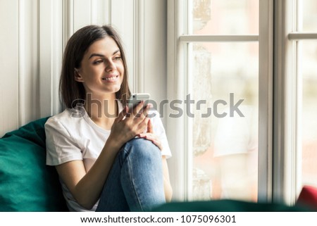 Beautiful smiling brunette girl wears white t-shirt using smartphone sitting on the windwsill