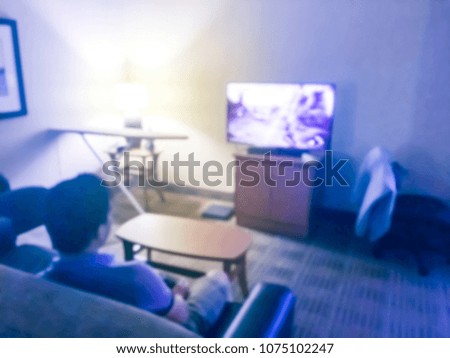 Abstract blurred rear view of young Asian man sitting on sofa and playing video game with joystick in front of big flat screen. Happy gamer playing indoor game at hotel while travelling