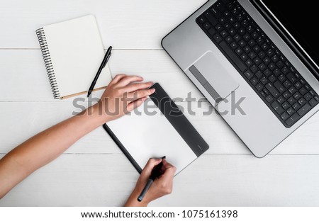 Top view of woman hands with graphic tablet drawing and retouching image on laptop computer, using digital tablet and stylus pen, free space. Graphic designer working on digital tablet. Blank notebook