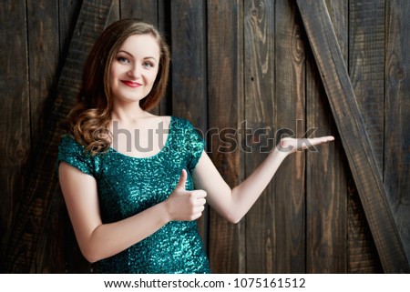 Beautiful smiling young woman in emerald dress pointing with hand on wood background and showing thumbs up gesture. St. Patricks Day, March 17. Girl showing empty copy space. Proposing product