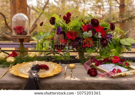 A red rosebud lying on a gold platter with a black veil, next to a glass of red wine, golden spoon and knife, golden fruits and flowers on an old wooden table. Decoration, gothic wedding. Copy space.