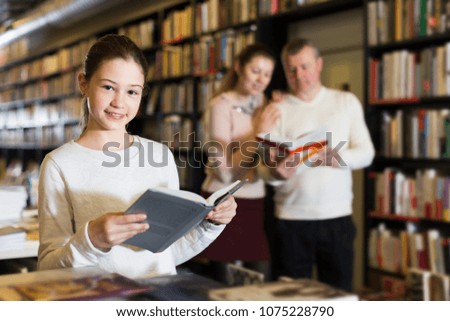 Smiling tweenager girl reading interesting book in bookshop during shopping with parents