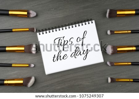 Makeup brushes arranged creatively on a dark gray textured background with "Tips of the day" text on a spiral notebook. Beauty concept. Top view or flatlay. 
