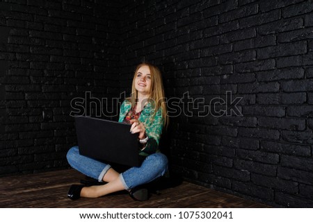 Stylish blonde girl in jacket and jeans with laptop against brick black wall at studio.