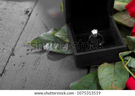 Solitaire Diamond Wedding ring in black velvet box with green rose leaves, red ribbons and marry me conceptual text on rustic black wooden background. Purpose and engagement concept. 
