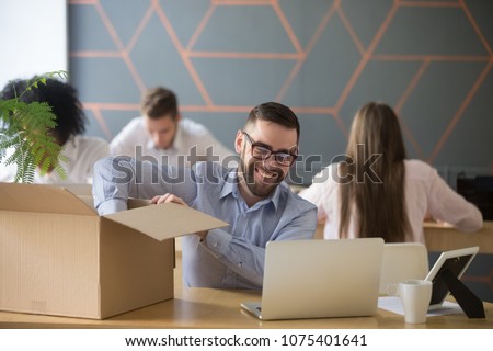 Smiling new male employee unpacking box with belongings at workplace, happy hired office worker newcomer on first working day concept, excited millennial businessman put laptop on desk in coworking Royalty-Free Stock Photo #1075401641