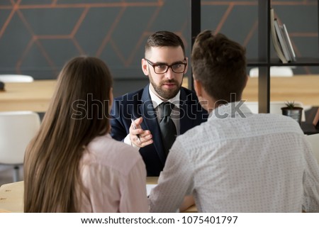 Serious investment broker, financial advisor or bank worker in suit and glasses consulting young couple giving legal advice, offering loan or presenting insurance services convincing to make deal Royalty-Free Stock Photo #1075401797