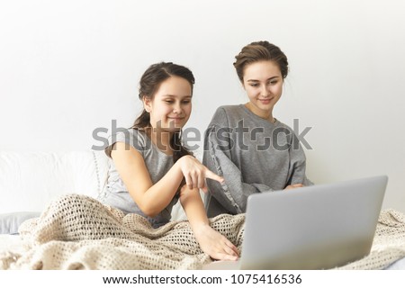 Picture of cheerful young woman dressed in gray sweatshirt sitting on bed with her little sister who is pointing index finger at laptop screen while watching movie or shopping online together