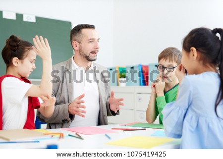 Group of Caucasian 1st graders sitting at desk with professional male teacher, drawing and communicating with each other Royalty-Free Stock Photo #1075419425