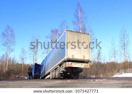 Tilted semi truck in roadside ditch – vehicle crash, skid, black ice, road accident on day winter road on trees and blue clear sky background. Sad driver call on the phone. Royalty-Free Stock Photo #1075427771