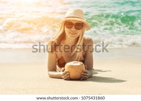 Portrait of smiling young woman in sunglasses and summer hat laying on sea beach, drink coconut and looking on copy space
