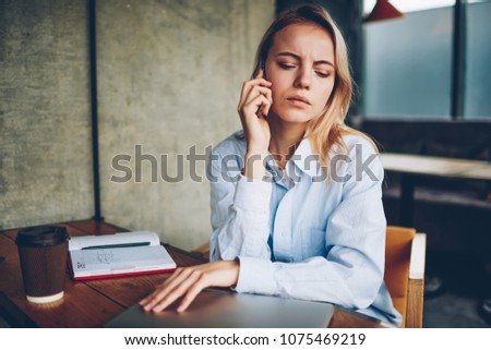 Serious hipster student calling to operator on smartphone device while sitting in coffee shop.Pensive young woman attentively listening audio message from operator on telephone resting in cafe