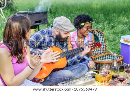 Happy friends playing guitar at picnic lunch in a park outdoor - Young people having fun and laughing together at barbecue in garden - Friendship, bbq and youth lifestyle concept