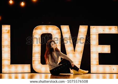 Full length portrait of a beautiful caucasian girl sitting on the floor smiling against a black wall decorated with lighting word LOVE.