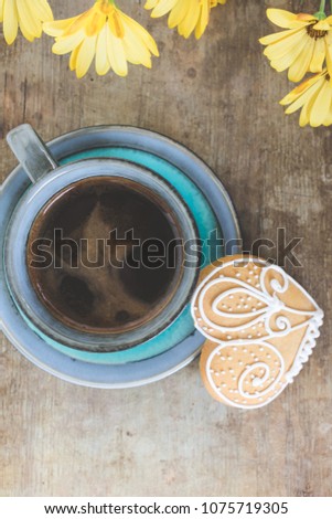 Top view of a vintage cup filled with black coffee with a heart shaped gingerbread cookie on old rustic wooden background with blooming spring flowers. Spring or summer breakfast