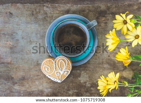 Top view of a vintage cup filled with black coffee with a heart shaped gingerbread cookie on old rustic wooden background with blooming spring flowers. Spring or summer breakfast