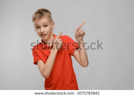 Portrait of blonde caucasian serious little boy in orange t-shirt showing on blank area on gray background