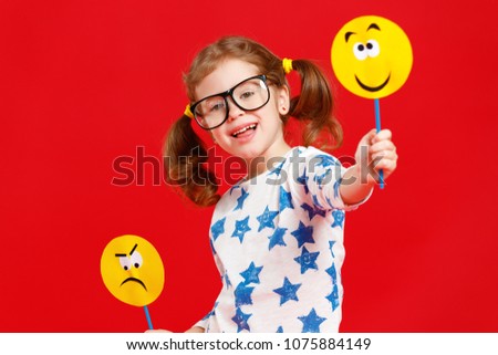 concept of children's emotions. child girl chooses between a sad and joyful smile on  colored red background
