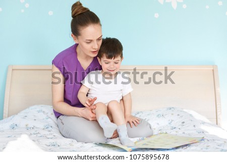 Indoor shot of affectionate young mother holds and embraces her small son, look attentively at book with colourful pictures, read interesting stories for children, sit on soft comfortable bed