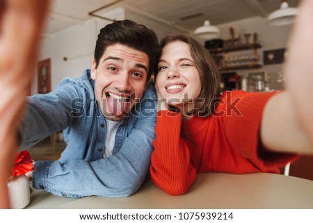 Caucasian couple woman and man smiling and showing tongue at camera while taking selfie in cafe