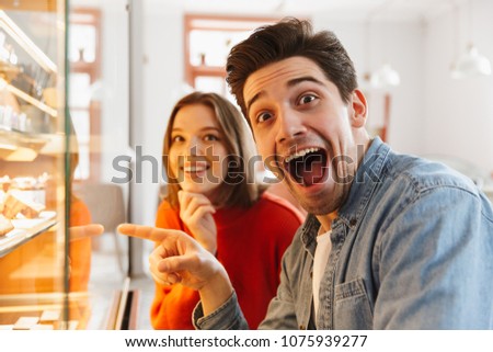Joyful couple woman and man in casual wear rejoicing while choosing sweets and cakes in cozy bakery