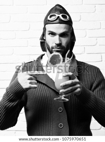 handsome bearded aviator man with long beard on serious face holding glass of alcoholic cocktail in gray knitted sweater with hat and glasses on white brick wall background
