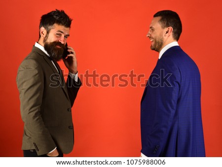 Men with beards stand face to face on red background. Machos in classic suits talk on mobile phone. Businessman with smiling face holds cell phone. Business talk and communication concept.