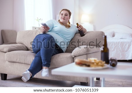 Bad nutrition. Stout red-head woman drinking beer while watching television Royalty-Free Stock Photo #1076051339