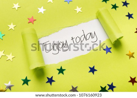 Inspirational phrase Enjoy today appearing behind torn yellow paper with small glittering stars. Quote for banner, poster or clothing design, copy space